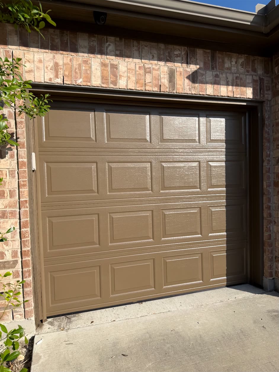 Almond-coloured short-panel single garage door closed on a brick home, photographed in bright afternoon sunlight.
