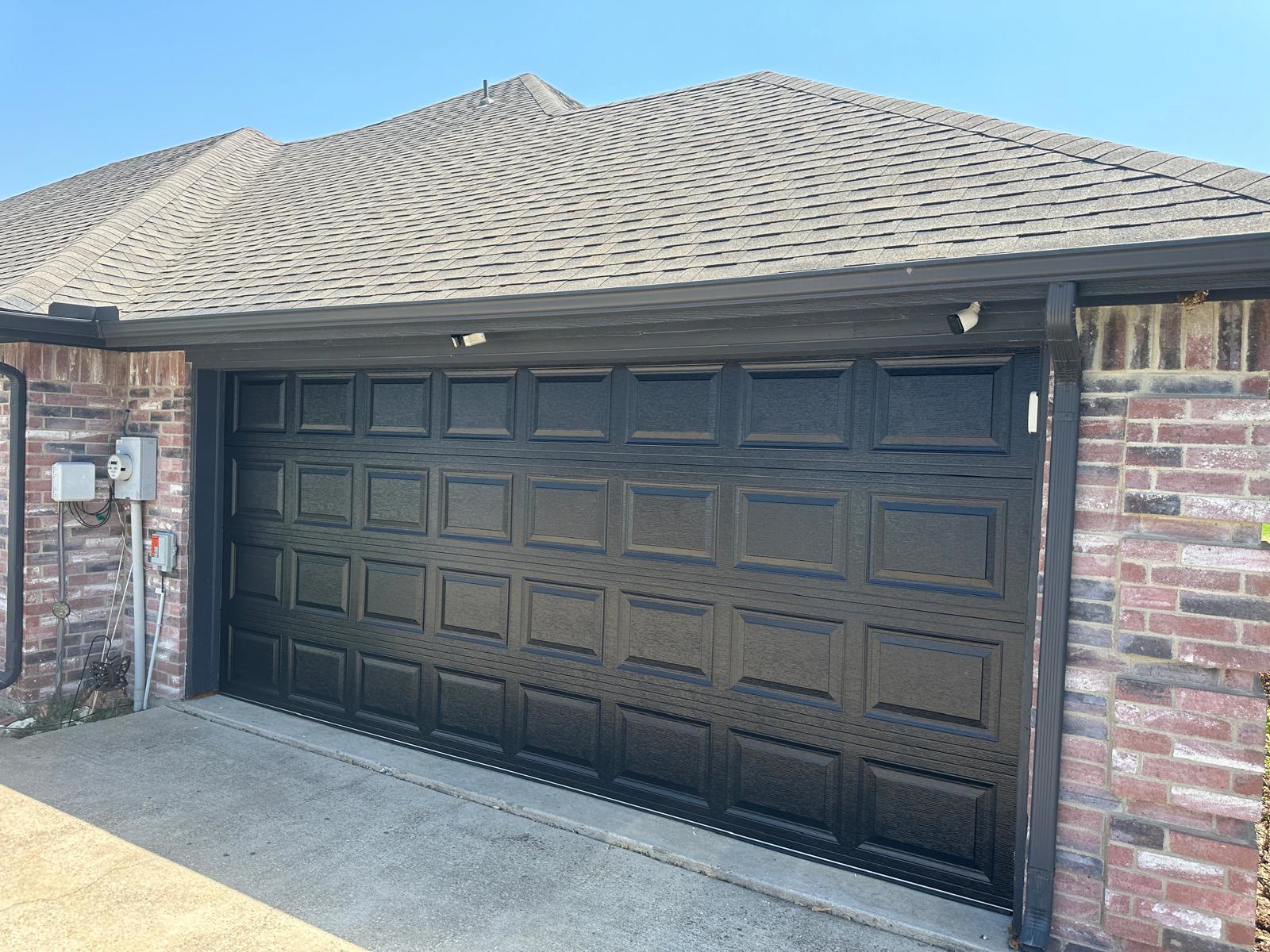 Black short-panel double garage door closed on a red brick single-storey home under a clear blue sky, with the electrical meter and disconnect box on the wall to the left.