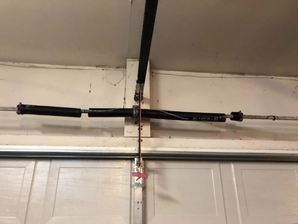 Broken torsion spring above a white sectional garage door: the coils have snapped and pulled apart, leaving a clear gap of bare winding shaft between the two halves of the spring.