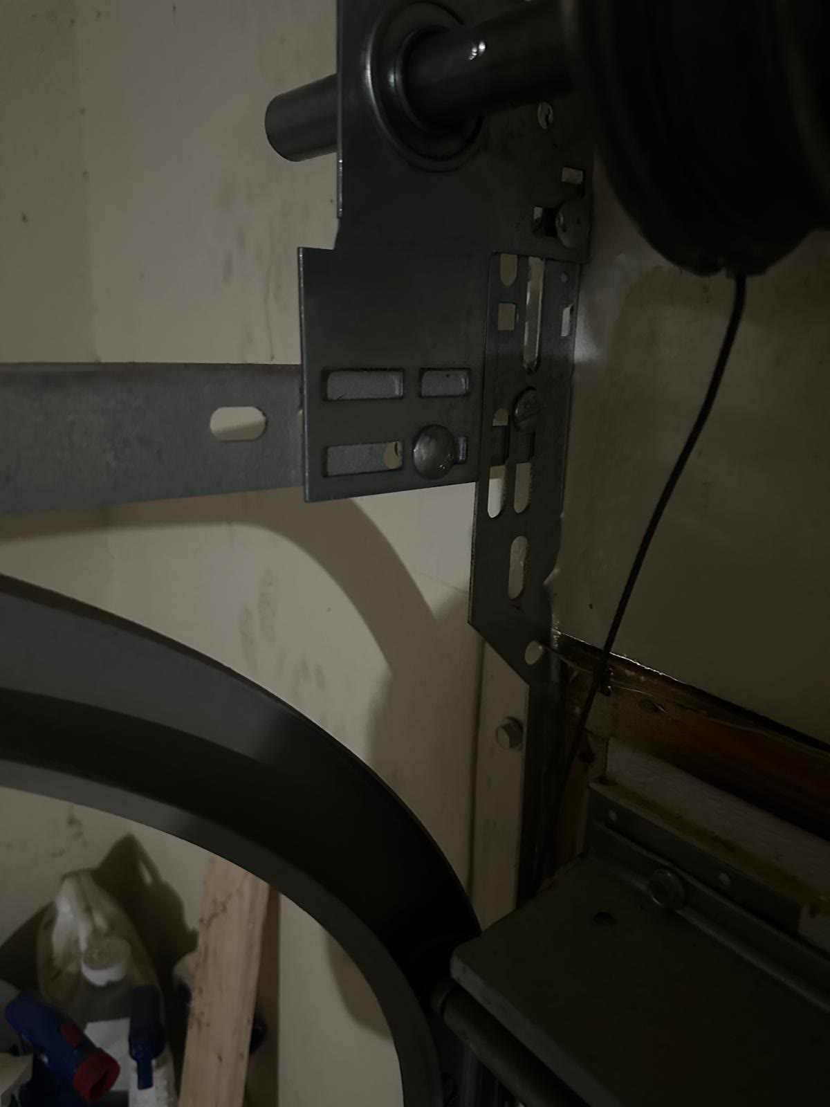 Close-up inside a dim garage of the end bearing plate and grooved cable drum at the top of a door track, with the lift cable running down from the drum past the curved section of track.