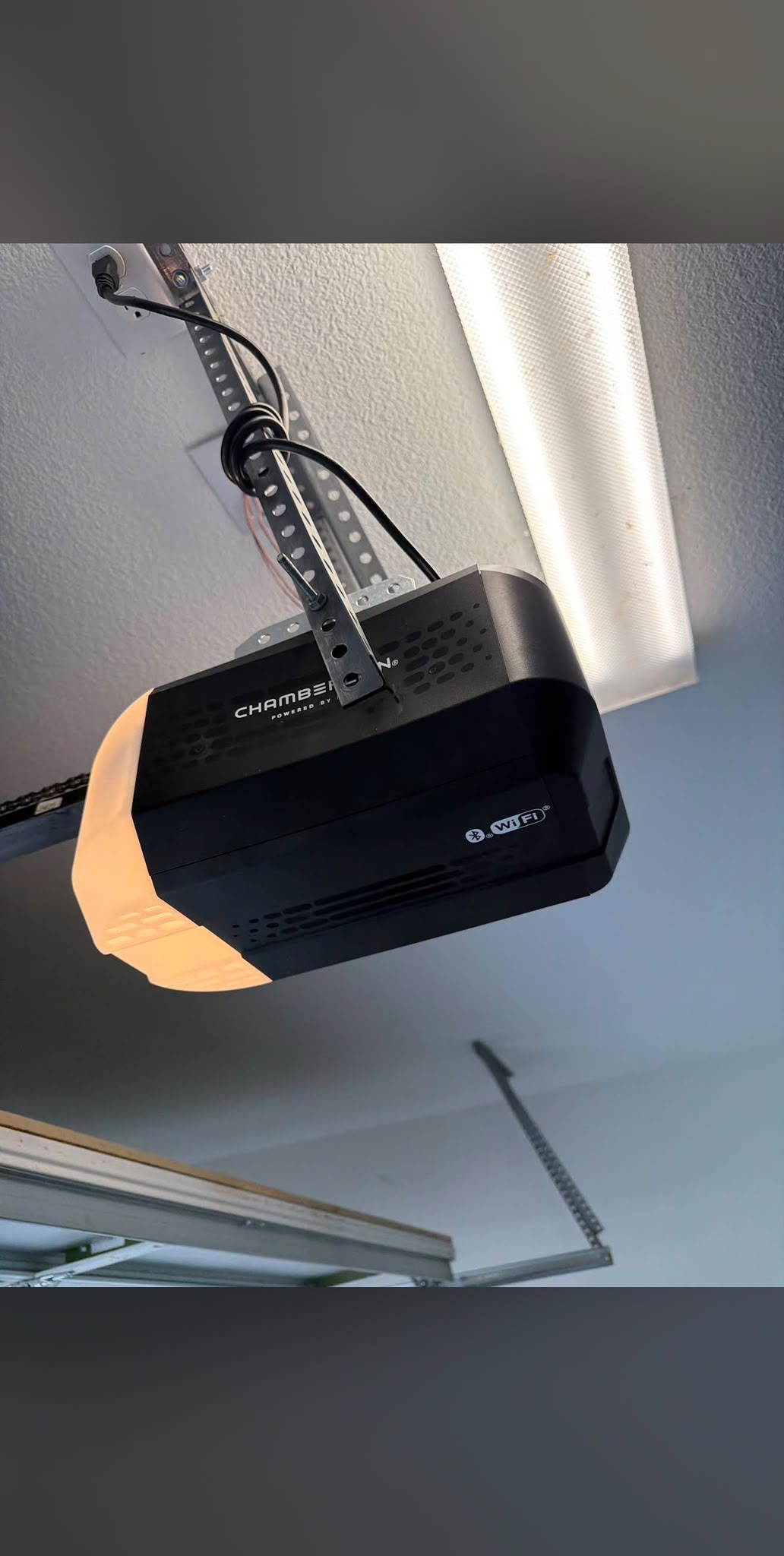 Close-up of a black Chamberlain Wi-Fi garage door opener newly mounted to a garage ceiling, its power cord looped up to the outlet and the drive rail running out of frame.