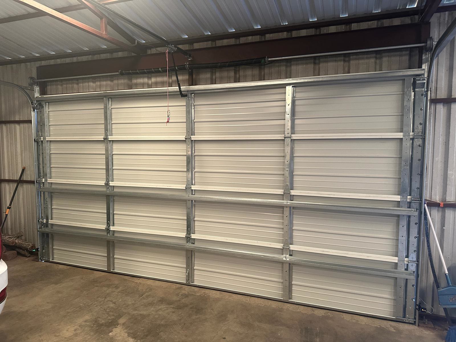 Newly installed white ribbed sectional door seen from inside a steel building, its galvanised struts, torsion spring, cable drums and red emergency release cord all visible against the metal wall.