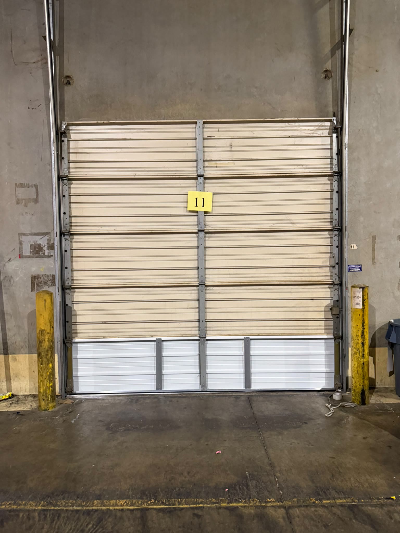 Commercial sectional door in a warehouse bay after its damaged bottom sections were replaced, the bright new white steel panels contrasting with the older cream panels above.