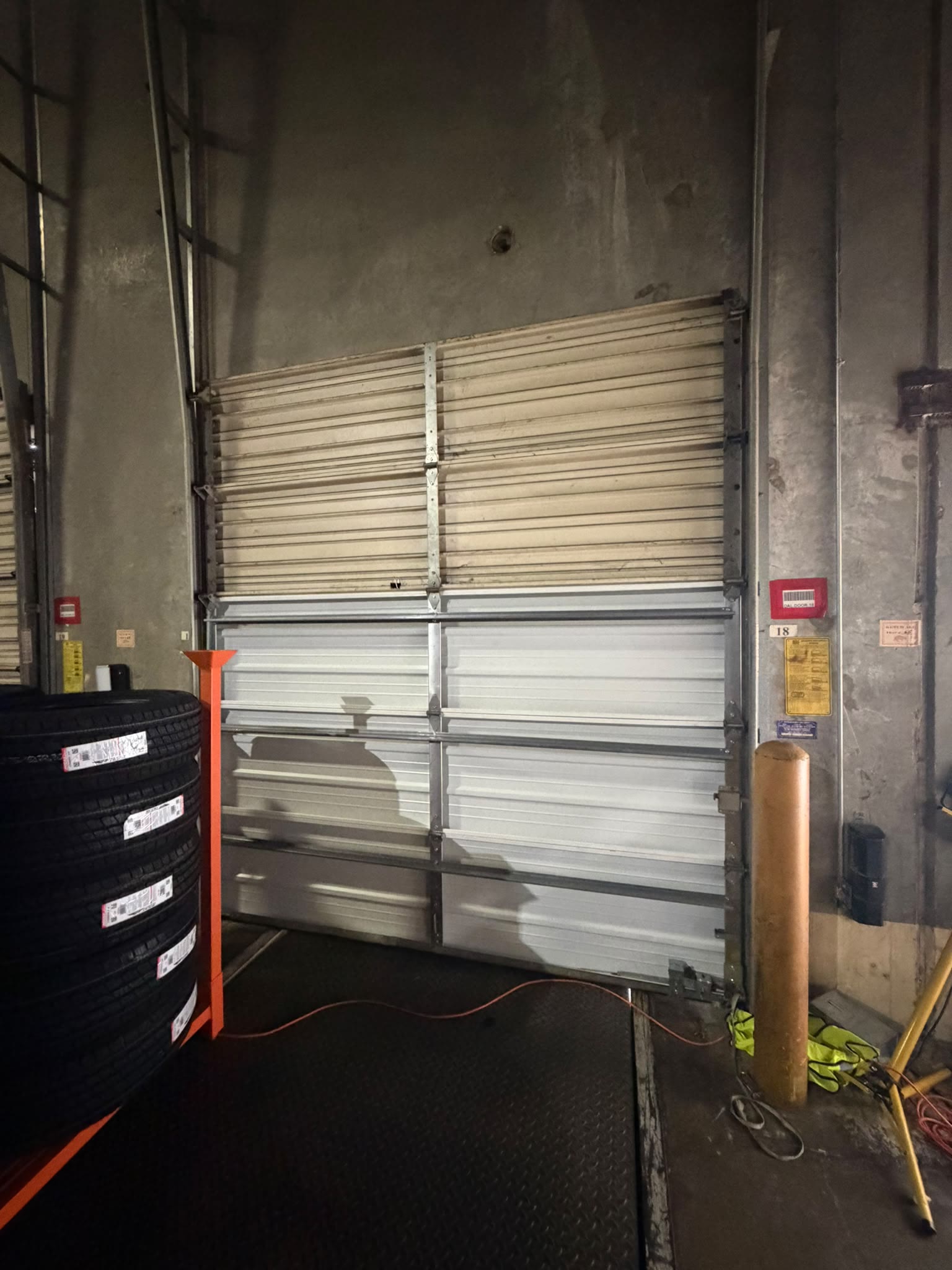 Commercial insulated sectional door at a warehouse loading bay, raised partway above the dock leveller, with stacked truck tyres and an extension cord in the foreground.