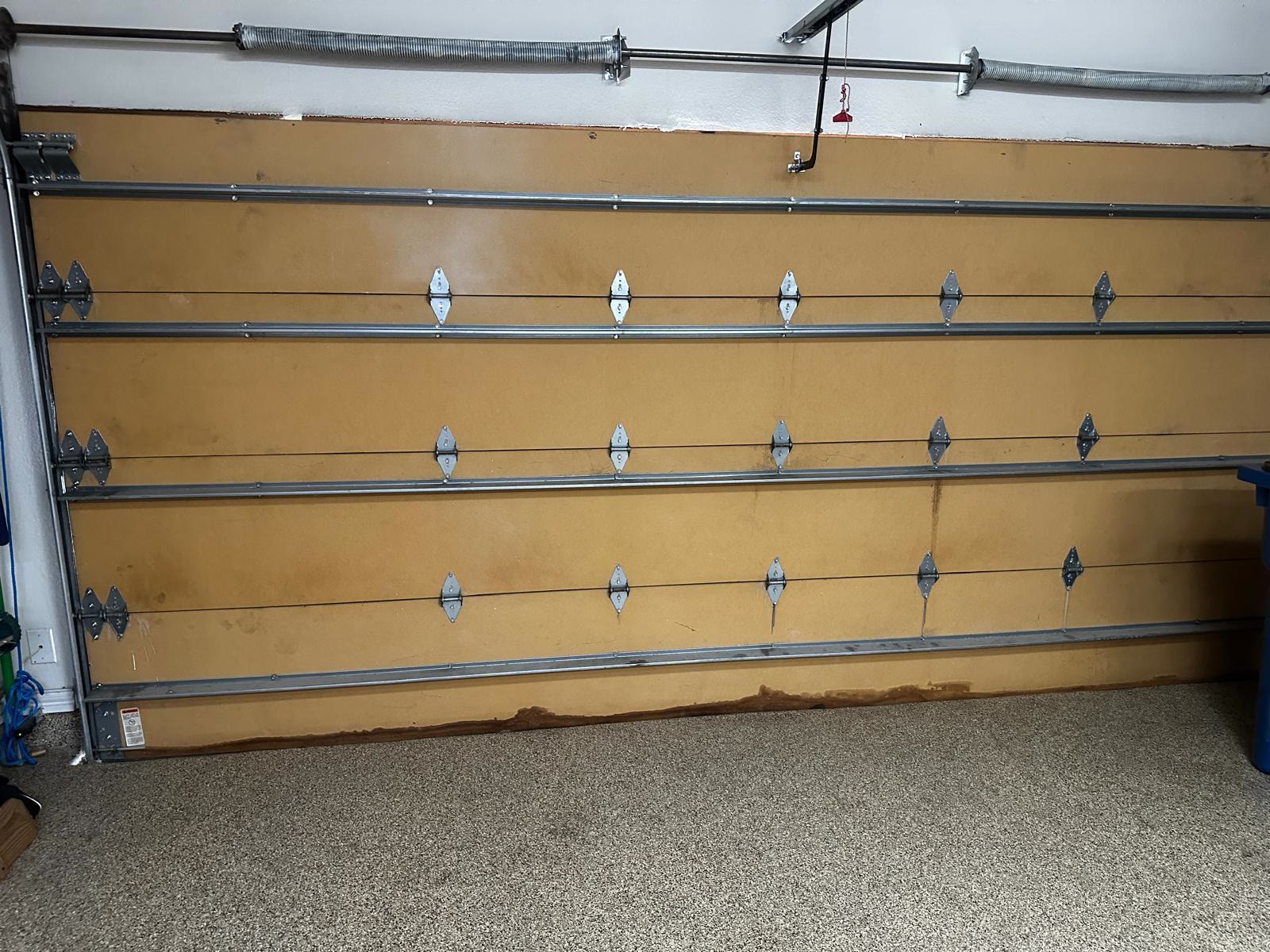 Inside face of a tan sectional garage door, showing the galvanised reinforcement struts bolted across each section, the hinges at every joint and the two torsion springs on the shaft above the door.