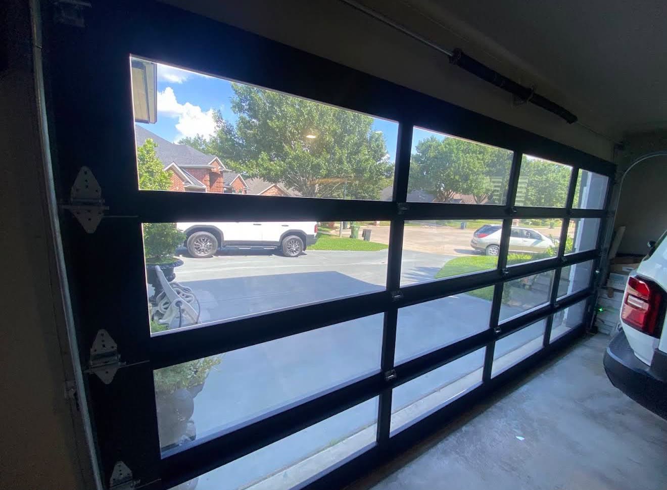 Full-view black-framed glass garage door seen from inside a residential garage, daylight spilling across the concrete floor with a white vehicle parked to the right.