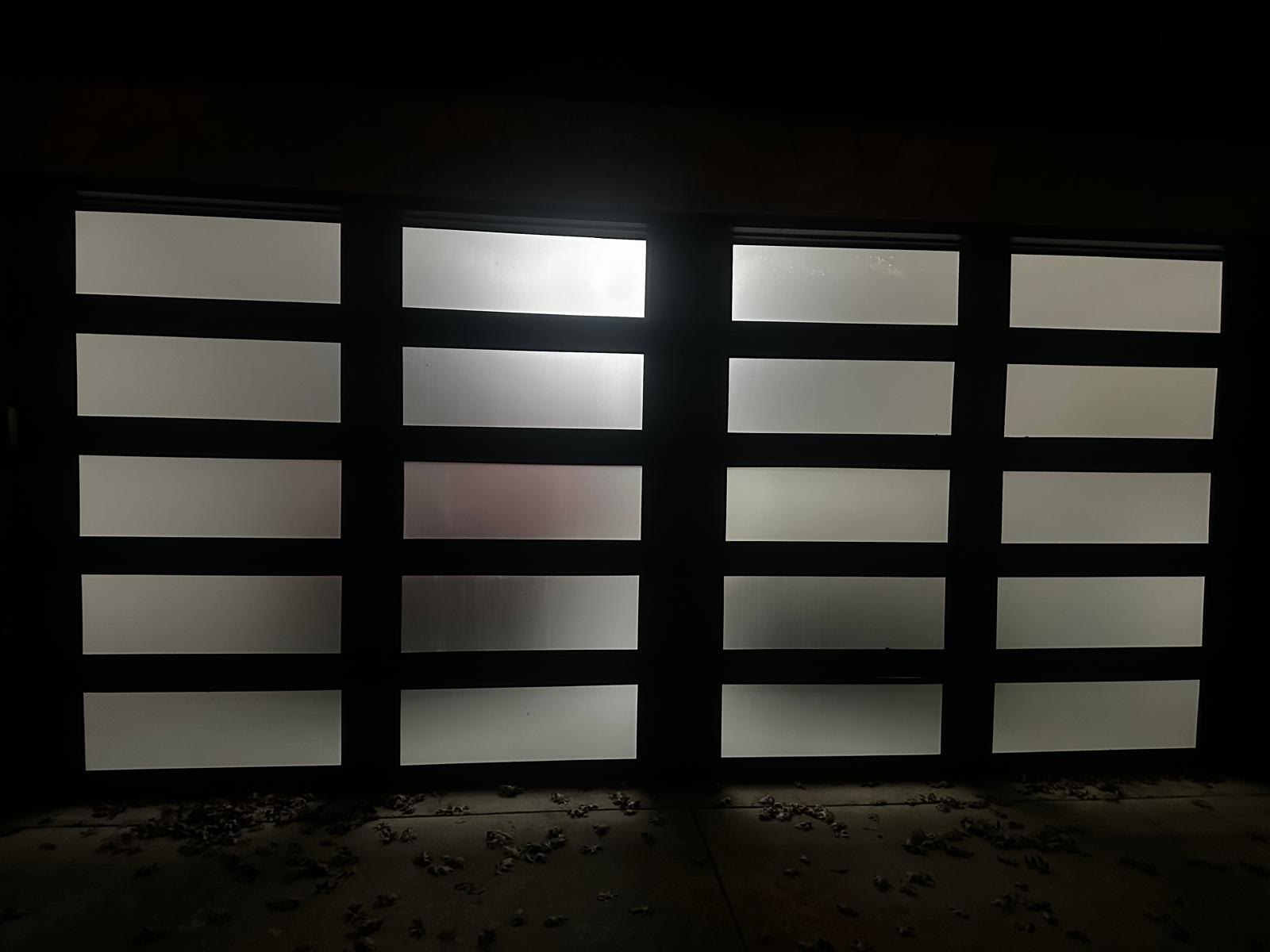 Modern full-view garage door with a black aluminium frame and frosted glass panels photographed from the driveway at night, the panels glowing softly from the light inside the garage.