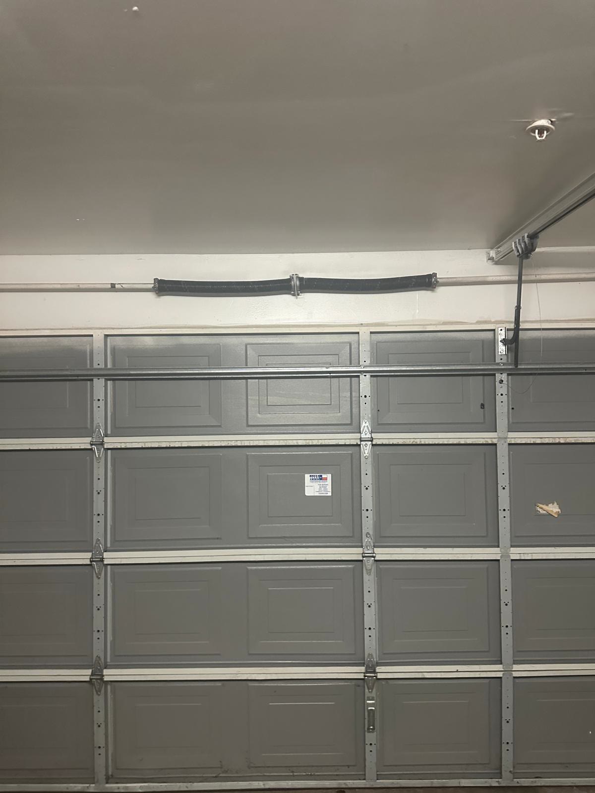 Grey raised-panel double garage door seen from inside the garage, closed, with its torsion spring and shaft mounted on the wall above the header and galvanised struts running across the sections.