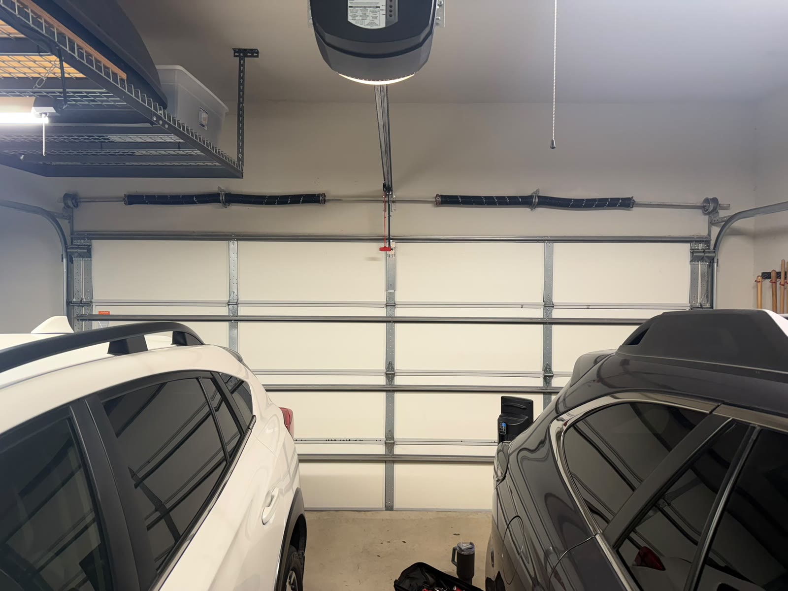 Two-car garage photographed from between the parked cars, with a white insulated sectional door closed, a pair of torsion springs on the shaft above it, an opener and its rail overhead and a ceiling storage rack to the left.