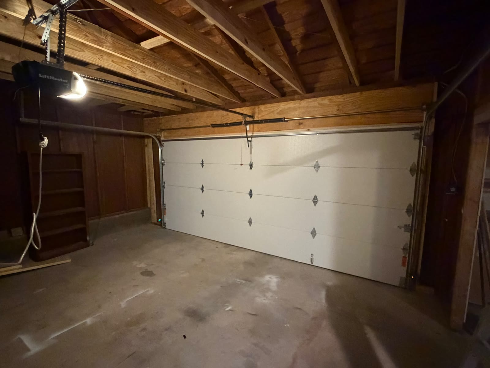 Residential garage interior with a white sectional door closed and a chain-drive opener mounted to the exposed wood ceiling framing, its light illuminating the empty concrete floor.