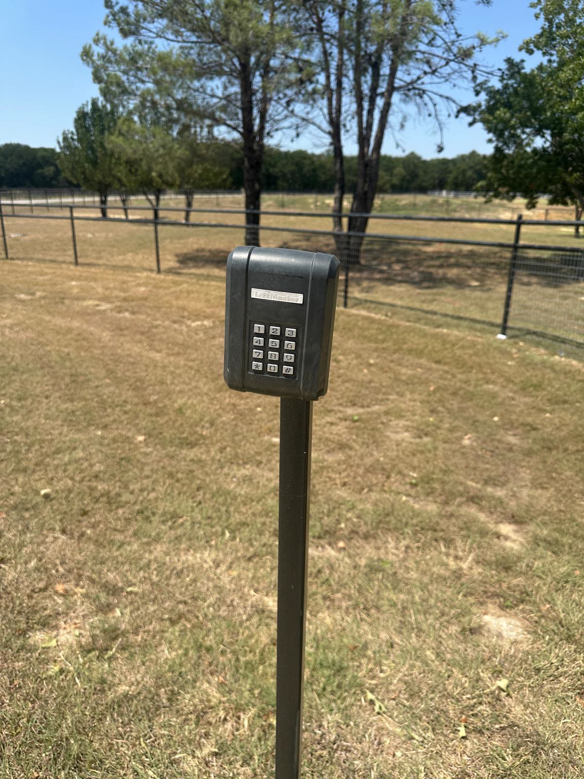 LiftMaster wireless entry keypad mounted on a slim steel post at the edge of a fenced pasture, with the tree line and paddock fencing behind it.