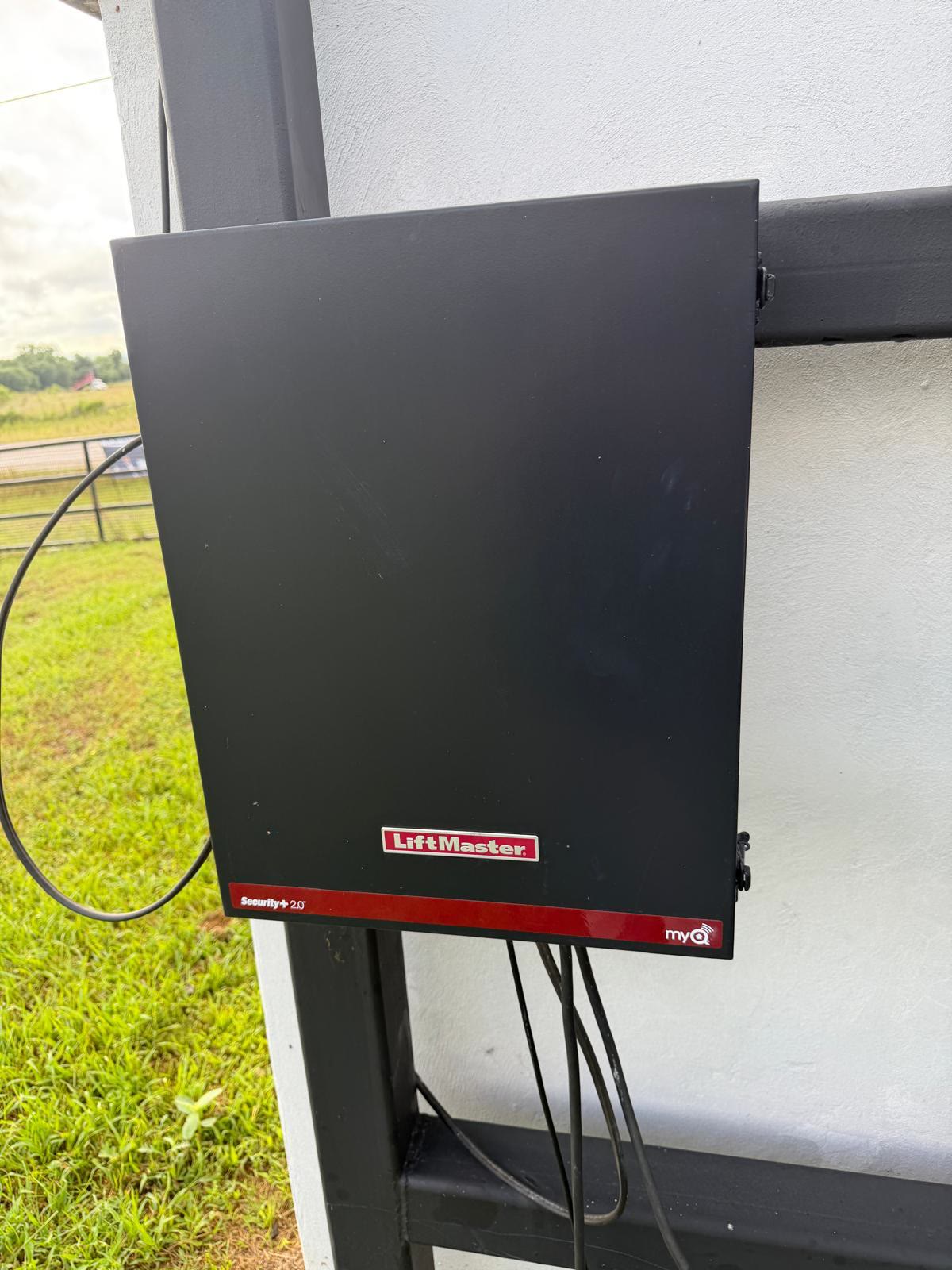 Black LiftMaster Security+ 2.0 control enclosure closed and bolted to a white gate post, its power and accessory cables looped out of the bottom, with green pasture behind.