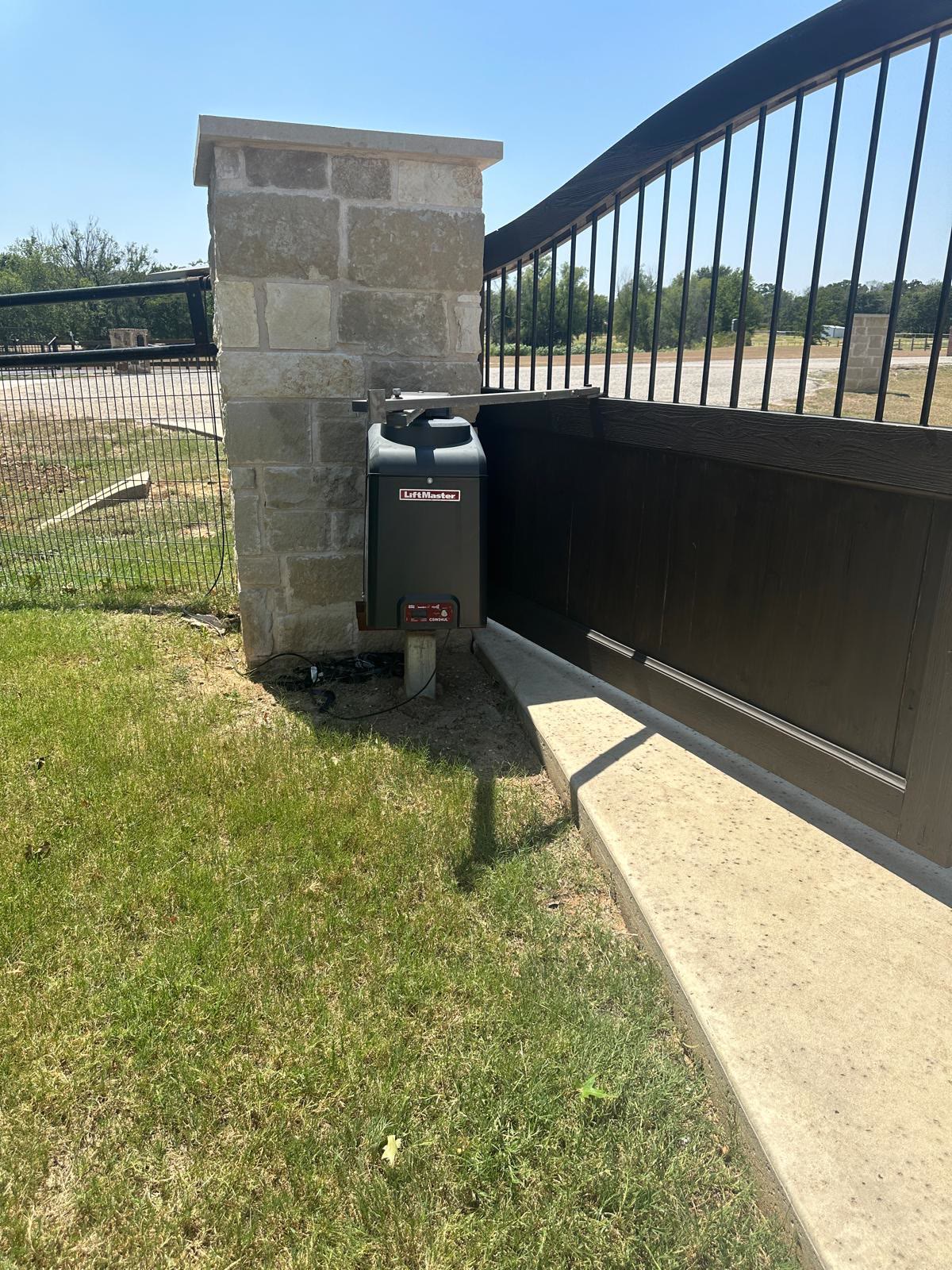 LiftMaster slide gate operator newly mounted on a concrete pad beside a stone gate column, driving a dark timber and wrought-iron driveway gate on a rural property.