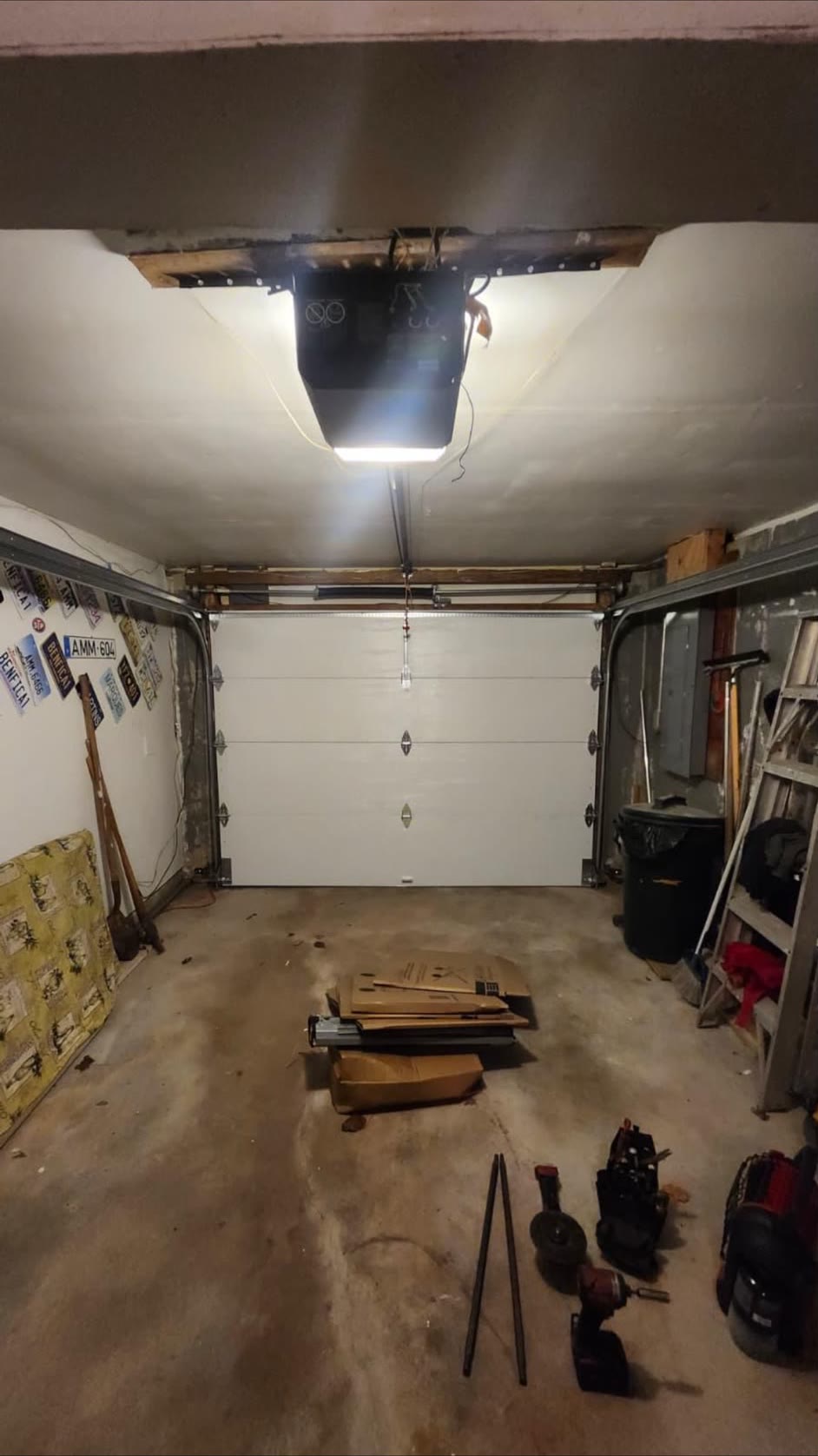 Single-car garage lit by a newly installed ceiling opener, with the opener packaging and cordless power tools still laid out on the floor in front of the white sectional door.