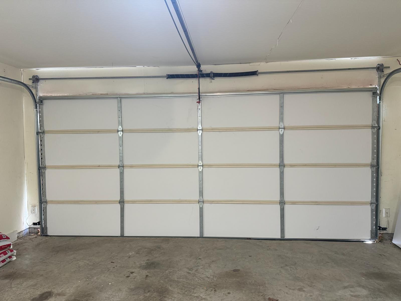 Newly installed white insulated sectional door filling an empty residential garage, its four galvanised struts, centre hinges and torsion spring visible against the pale wall.