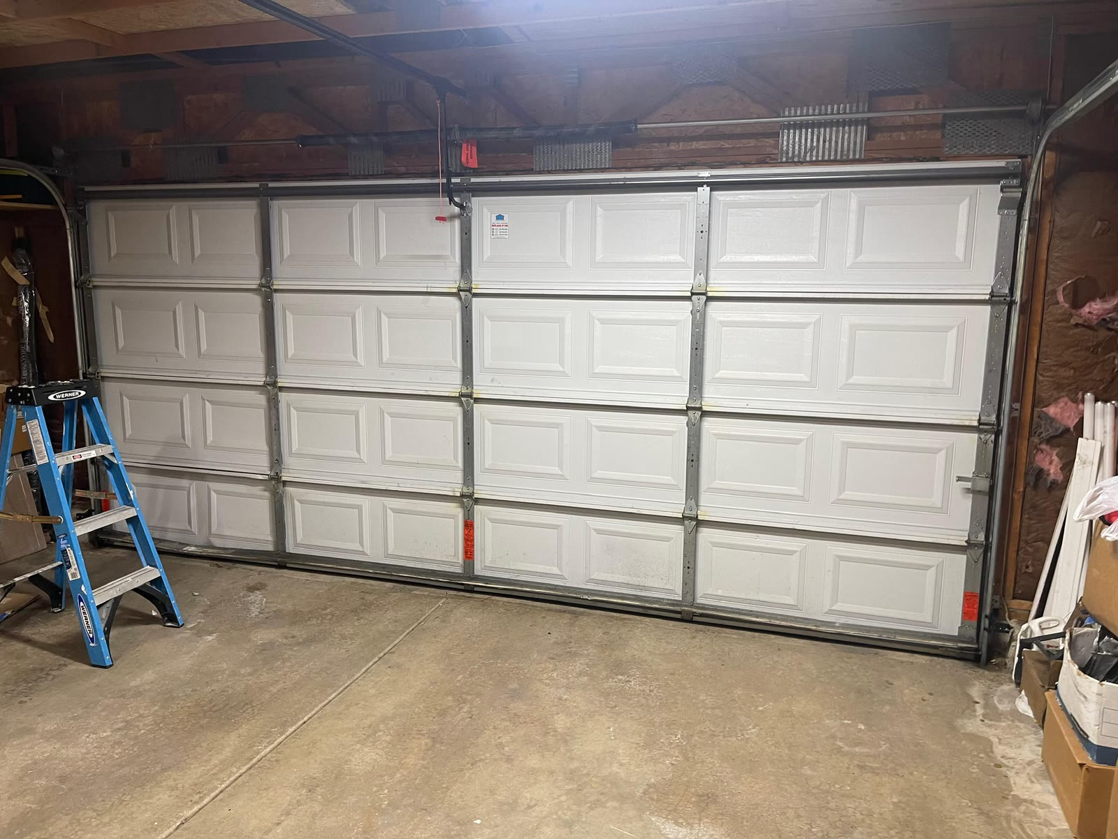 Newly installed white raised-panel double garage door seen from inside an unfinished garage, with a step ladder to the left and exposed wall insulation on the right.