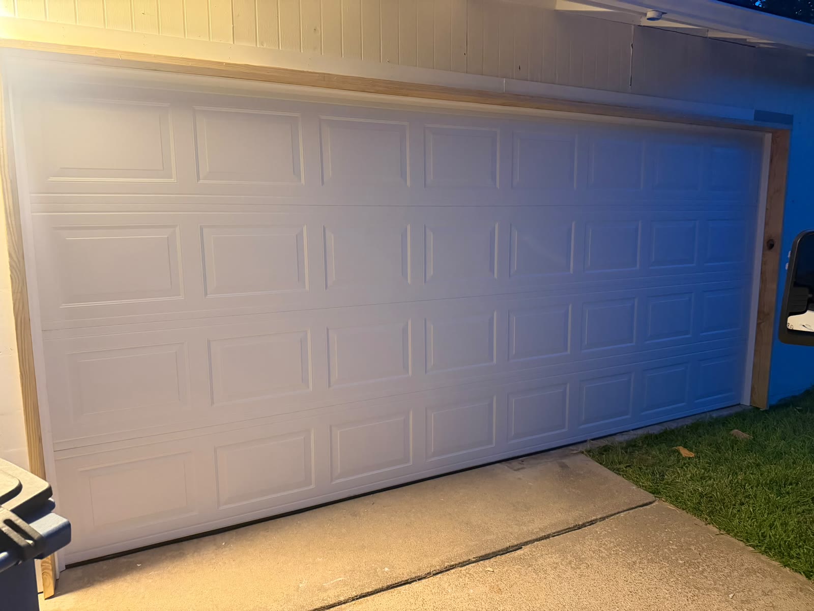 Newly installed white short-panel double garage door on a blue-sided house, closed and lit by the porch light at night.