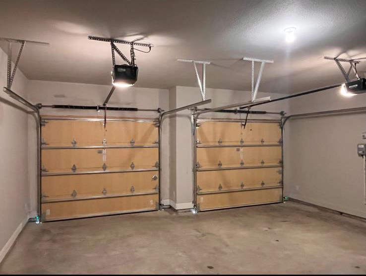Empty two-car garage after installation, with two new sectional doors backed in kraft-faced insulation, two chain-drive openers mounted and lit overhead and ceiling storage racks either side.