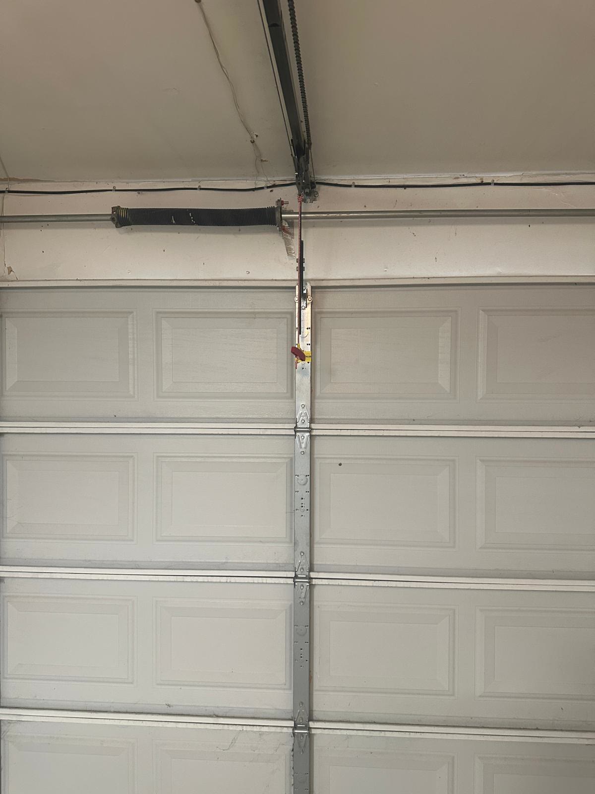 Single torsion spring on its shaft above a closed white raised-panel garage door, with the opener rail, chain, curved door arm and red emergency release cord running down the centre of the door.