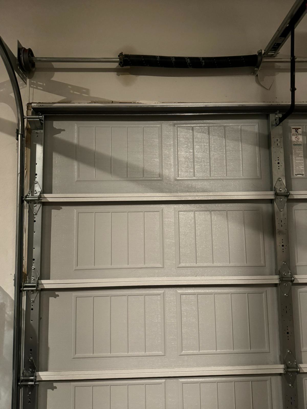 Inside a residential garage: a torsion spring on its shaft above the header, the cable drum and end bearing plate at the left-hand corner, and the hinges, brackets and vertical track running down the side of a closed cream short-panel door.