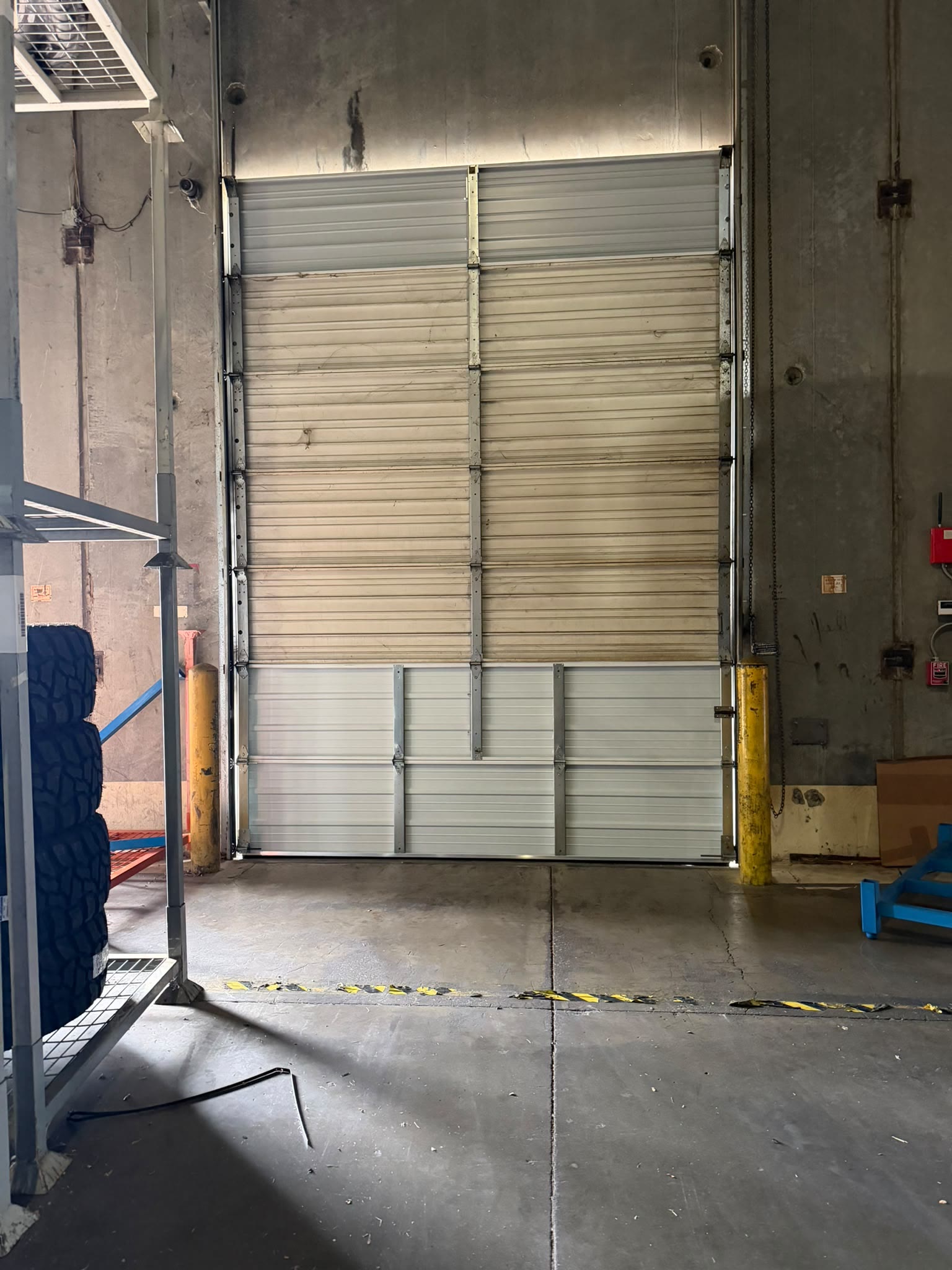 Tall commercial sectional garage door closed inside a concrete warehouse bay, flanked by yellow safety bollards with tyre racking to one side.