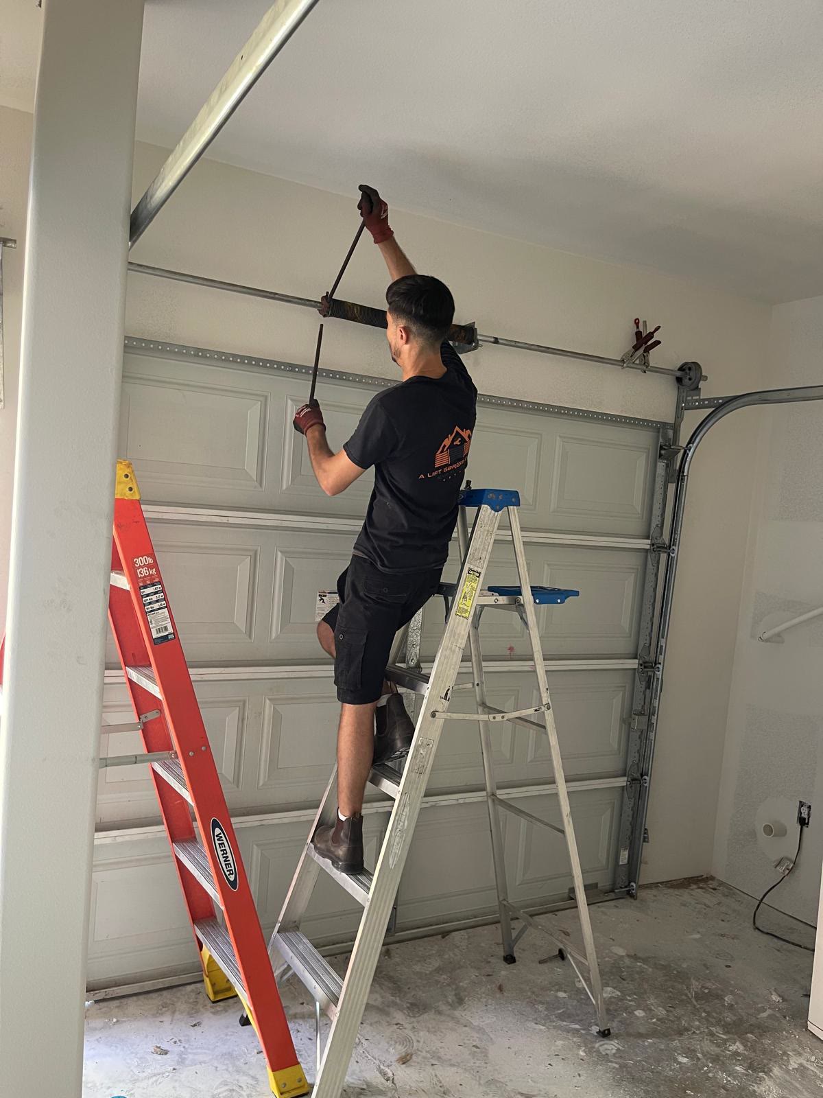 Technician standing on a step ladder and winding a torsion spring with a pair of winding bars, above the closed almond raised-panel door of an empty residential garage.