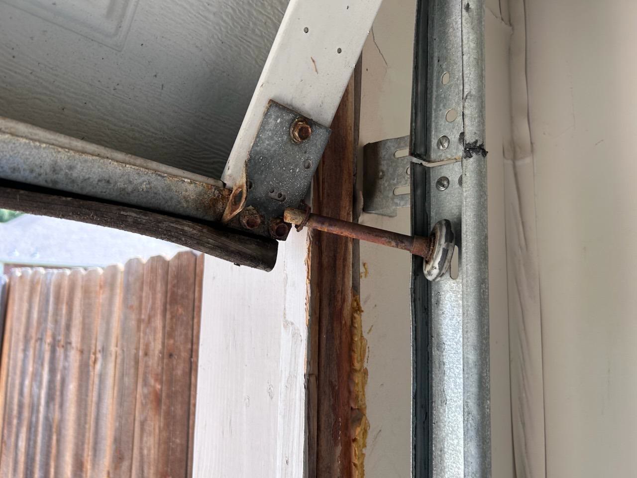 Close-up of a garage door that has come off its track: the top roller's rusted stem has pulled clear of the vertical track, the top section of the door has tipped away from the opening, and the timber jamb behind it is split and rotting.