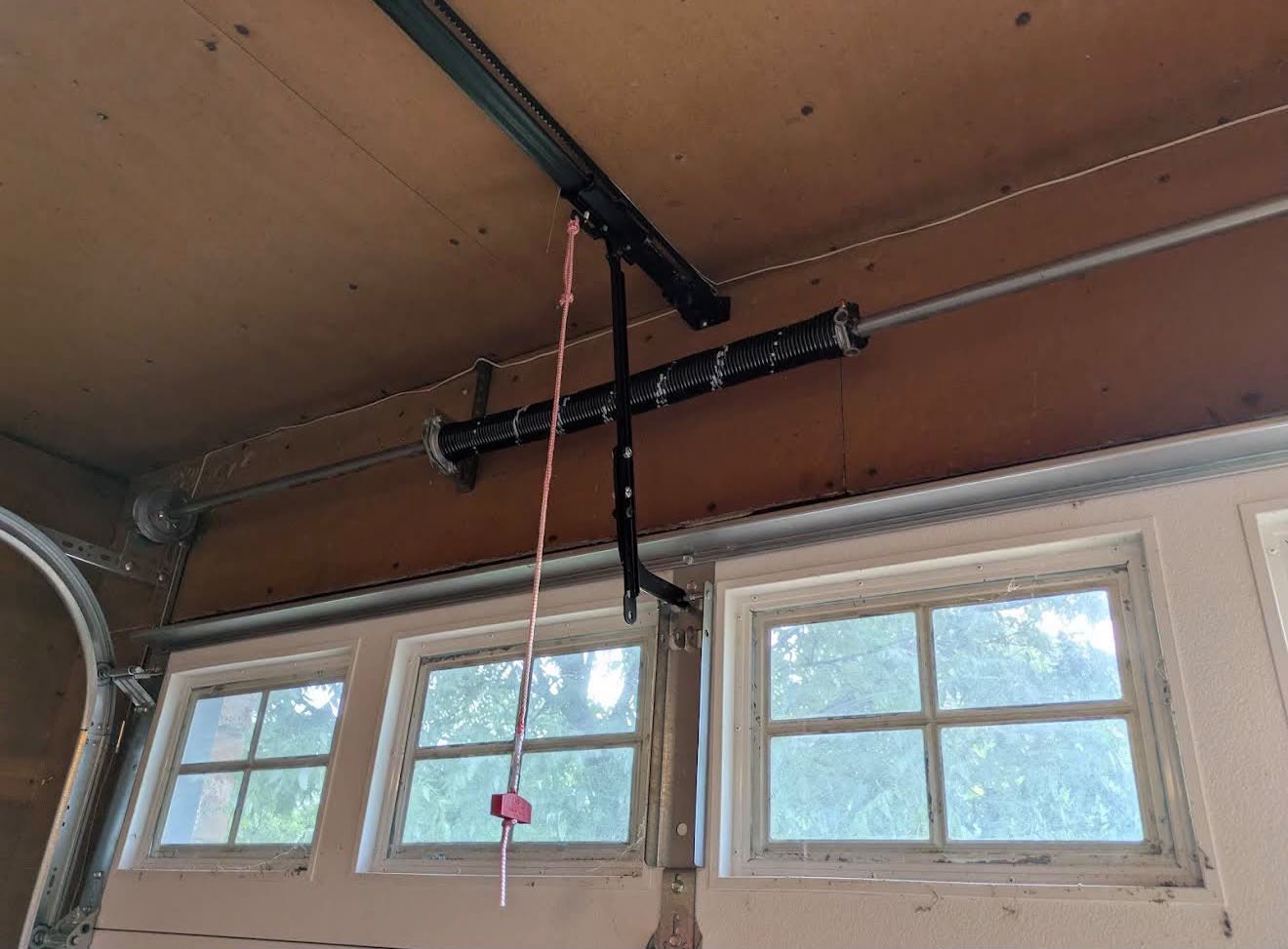 Torsion spring, winding cones and shaft mounted above the windowed top section of a garage door, with the opener rail, door arm and red release cord alongside, lit by daylight through the door glass.