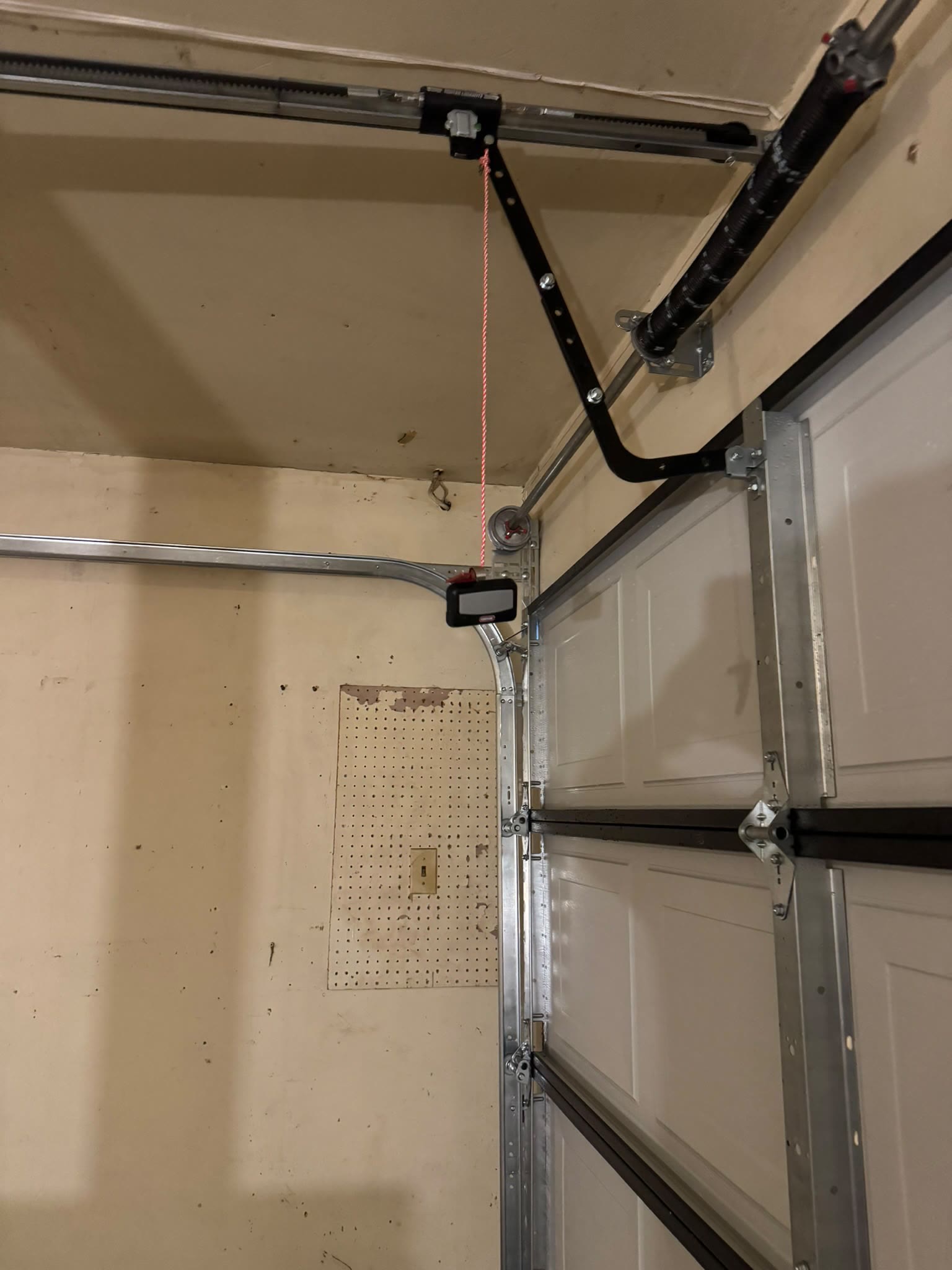 Corner of a garage ceiling showing the torsion spring on its shaft, the opener rail and curved door arm, the cable drum, and the wall control panel mounted beside the vertical door track.