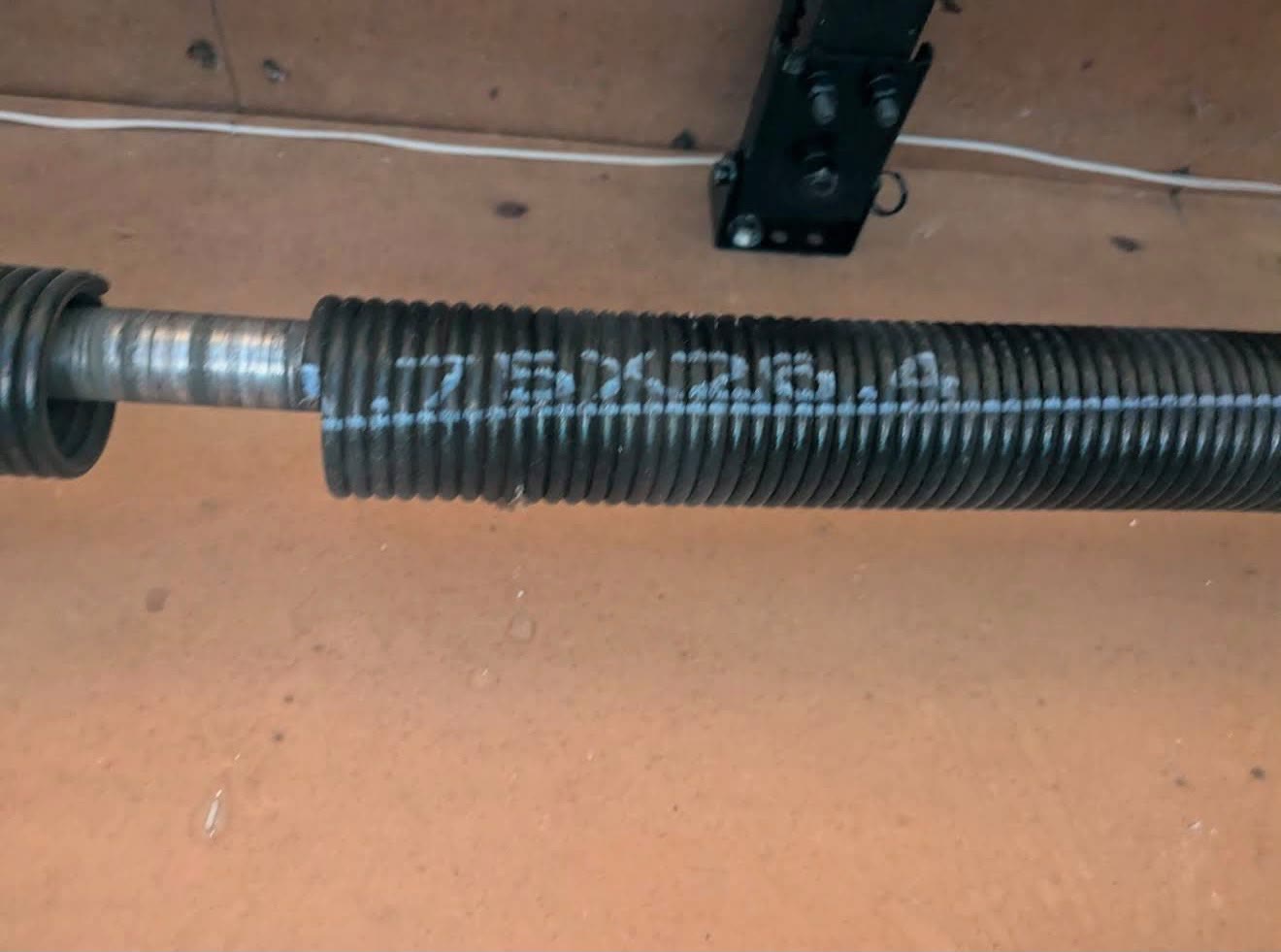 Close-up of a black torsion spring on its shaft with the wire gauge, inside diameter and length stencilled along the coils in white paint.