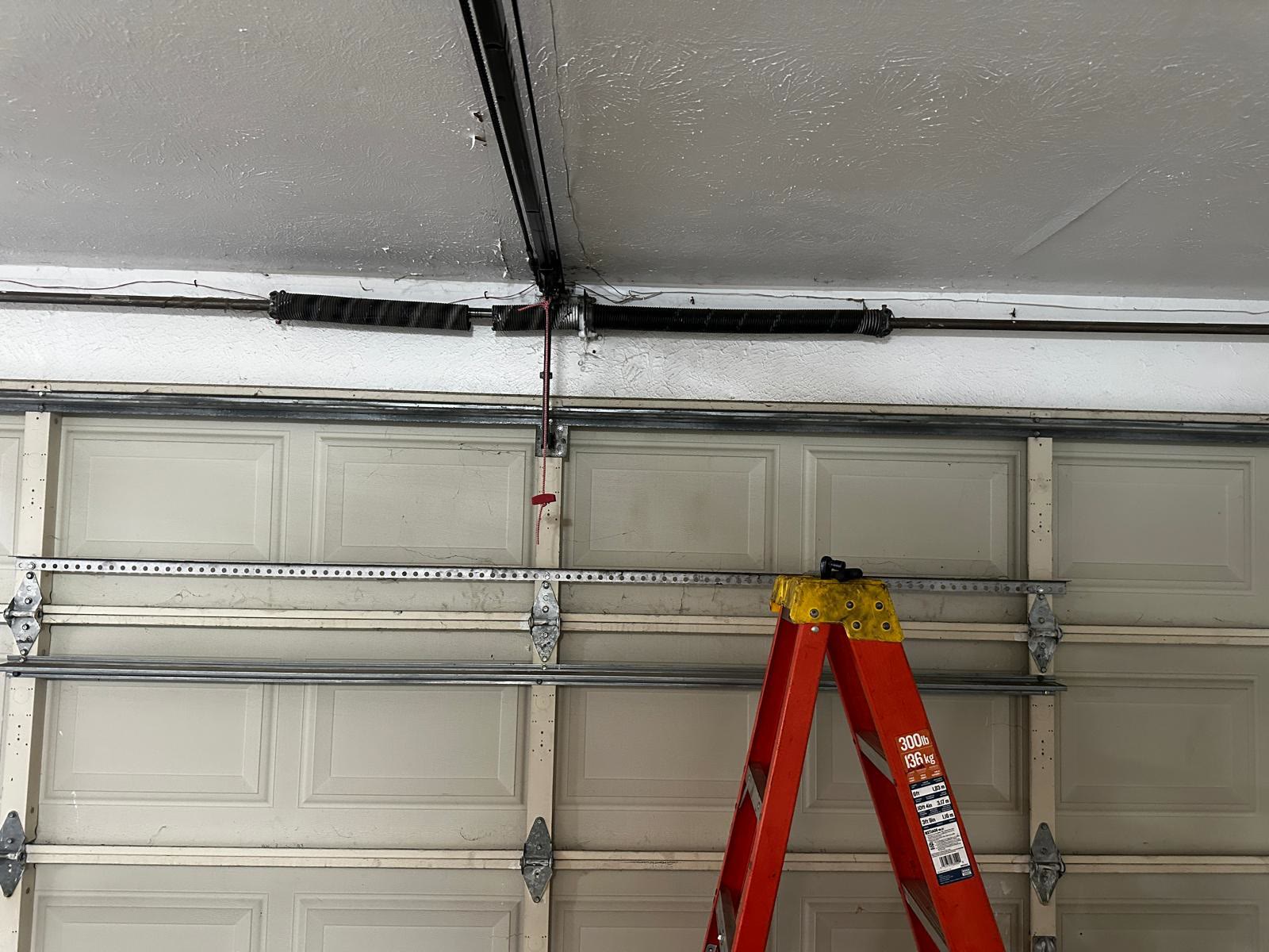 Two torsion springs mounted on the shaft above a closed almond raised-panel garage door, with the opener rail overhead, the red release cord hanging down and a fibreglass step ladder standing against the door.