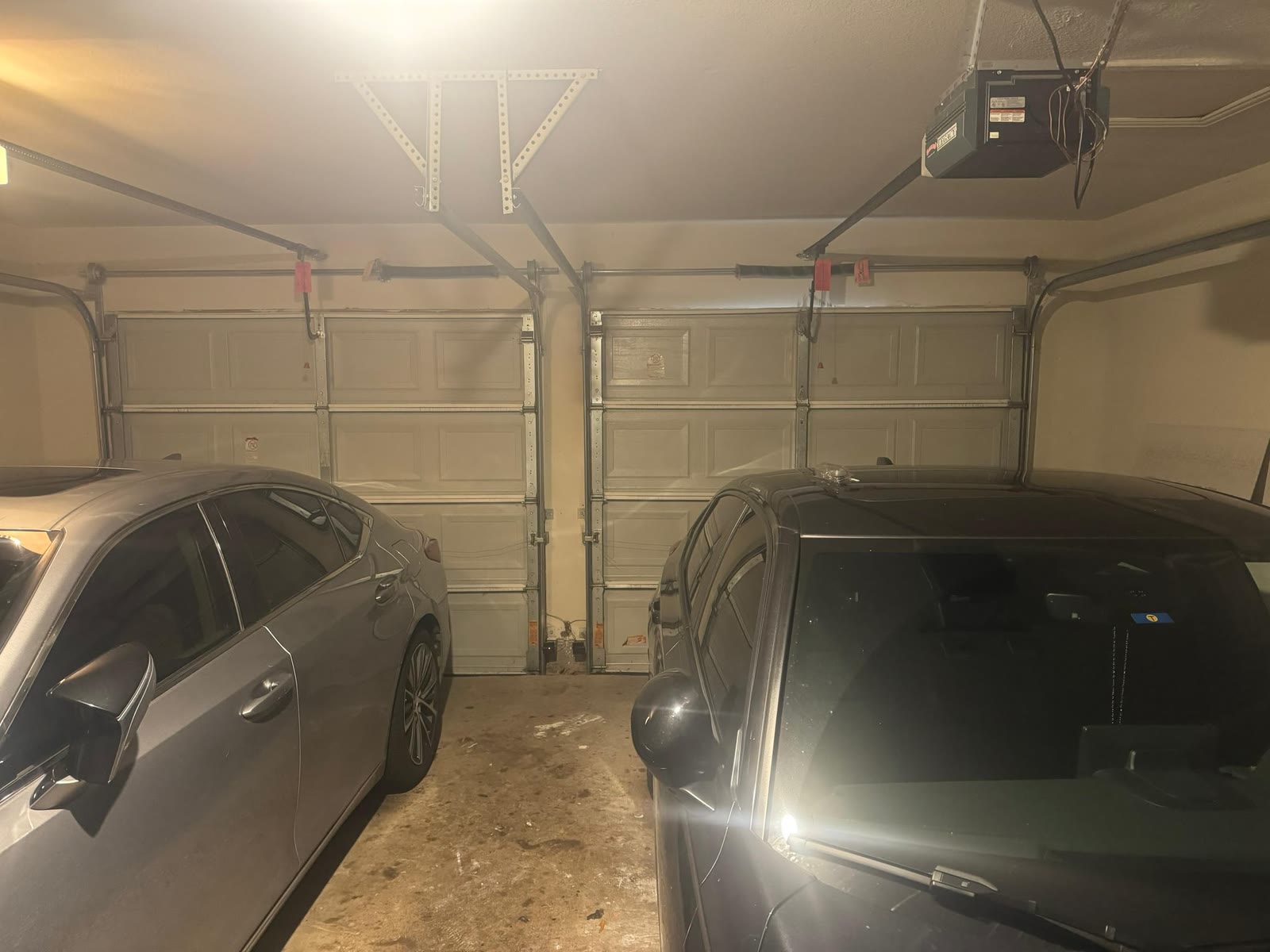 Two-car garage at night with a pair of single sectional doors, their torsion springs, safety cables and opener rails visible overhead, and two sedans parked either side.