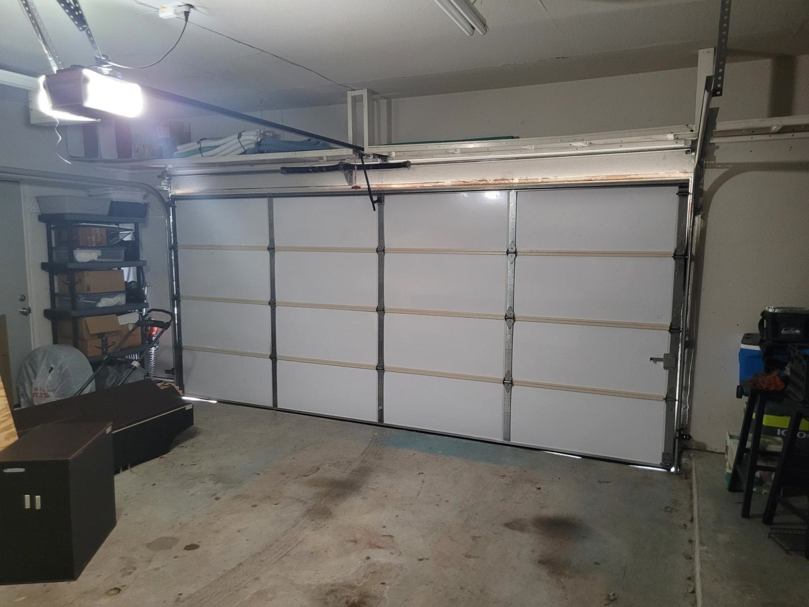 White insulated sectional door closed inside a lived-in residential garage, with a chain-drive opener lit up on the ceiling to the left and the door’s reinforcement struts running across the sections.