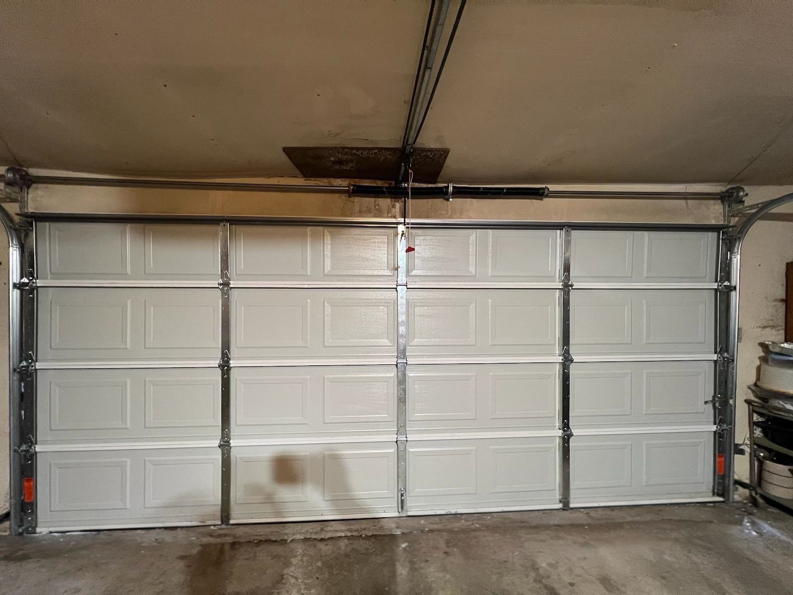 Newly hung white raised-panel double garage door seen from inside, closed and sealed to the floor, with its struts, hinges and torsion spring in place and the old timber header still exposed above.