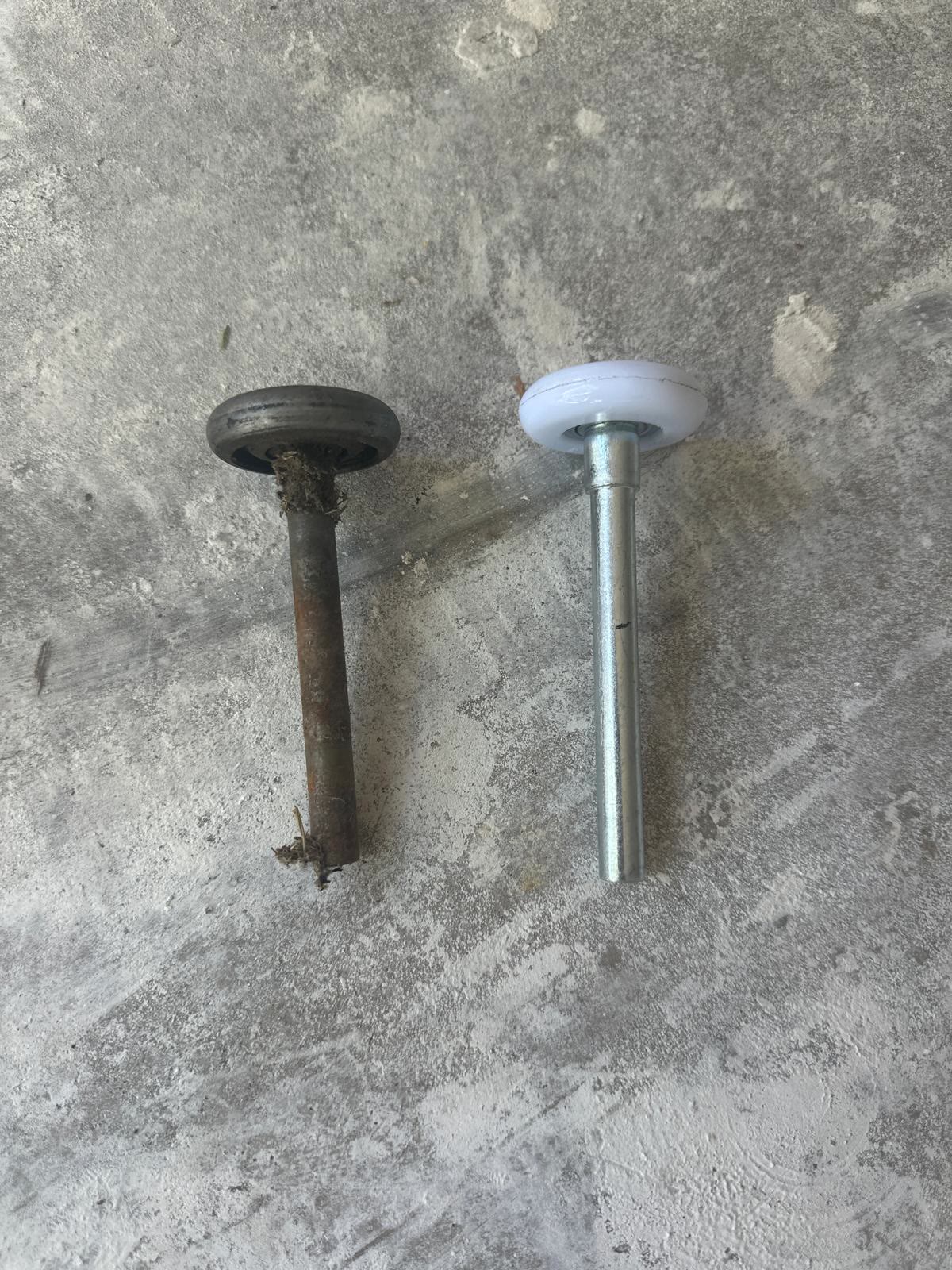 A worn steel garage door roller, its stem rusted and its bearing packed with grease and grit, laid on a concrete floor next to the clean white nylon roller that replaced it.